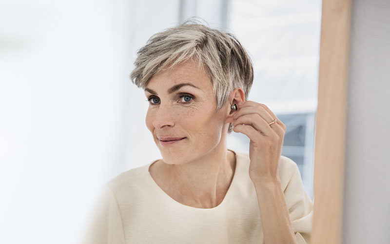 Older white woman putting an Oticon OPN invisible hearing aid in her ear