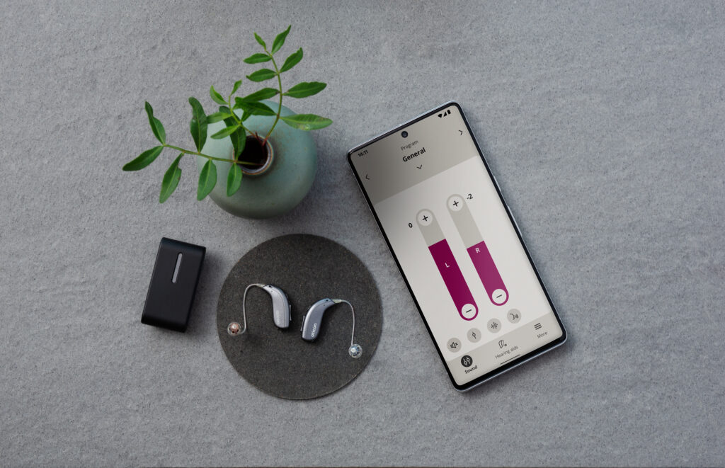 Oticon Real hearing aids next to a plant pot and a phone with the Oticon App open