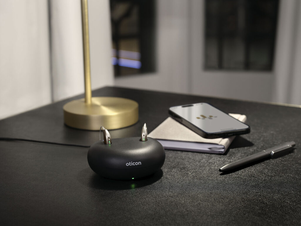 Oticon hearing aids in a charger on a desk. In the background there is a phone with the app open.