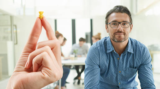 Small hearing aid held between the index finger and thumb with a man looking with people in the background